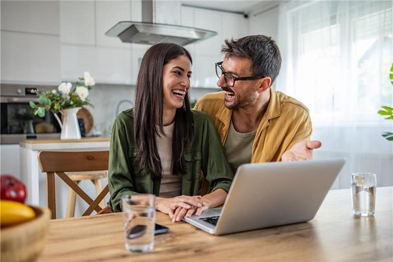 A couple laughing together at a kitchen table while using a laptop