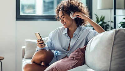 A woman wears over the ear headphones while scrolling on her phone and connecting to Optimum Internet.