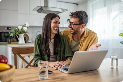 A couple laughing together at a kitchen table while using a laptop
